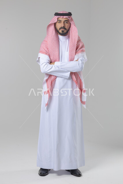 الاعتناء والاهتمام بالمظهر الخارجي، مفهوم الاناقة والرجولة، التقاط صور شخصية للوثائق الرسمية، بورتريه لشاب عربي خليجي سعودي يرتدي الثوب التقليدي والشماغ يقف بأيدي متقاطعة ينظر الى الكاميرا بتعابير السرور والثقة بالنفس، الجسم بالطول الكامل، خلفية بيضاء