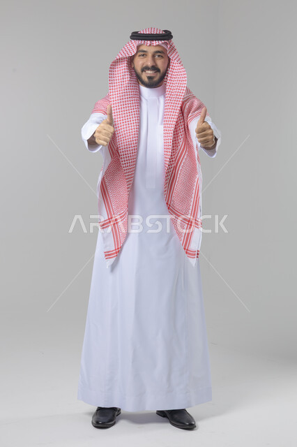  الإمتياز والتميز والنجاح، بورتريه لرجل عربي خليجي سعودي يرتدي الثوب التقليدي والشماغ ويرفع أصابع الإبهام لأعلى، إيماءات السعادة والإعجاب والموافقة، حركة أصبع تدل على القبول والرضا، الجسم بالطول الكامل، خلفية بيضاء 