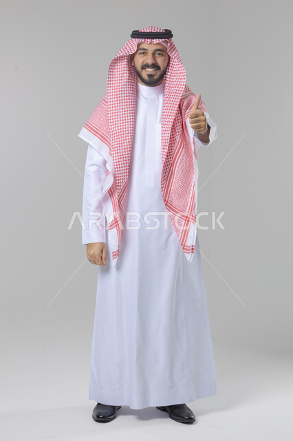  الإمتياز والتميز والنجاح، بورتريه لرجل عربي خليجي سعودي يرتدي الثوب التقليدي والشماغ ويرفع أصابع الإبهام لأعلى، إيماءات السعادة والإعجاب والموافقة، حركة أصبع تدل على القبول والرضا، الجسم بالطول الكامل، خلفية بيضاء 