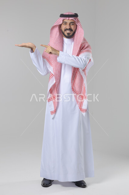 الإشارة إلى كف اليد, الوقوف باستقامة واعتدال, بورتريه لرجل عربي خليجي سعودي يرتدي الثوب التقليدي و الشماغ يحمل بيديه شيء ما, صورة للجسم بالطول الكامل, خلفية بيضاء