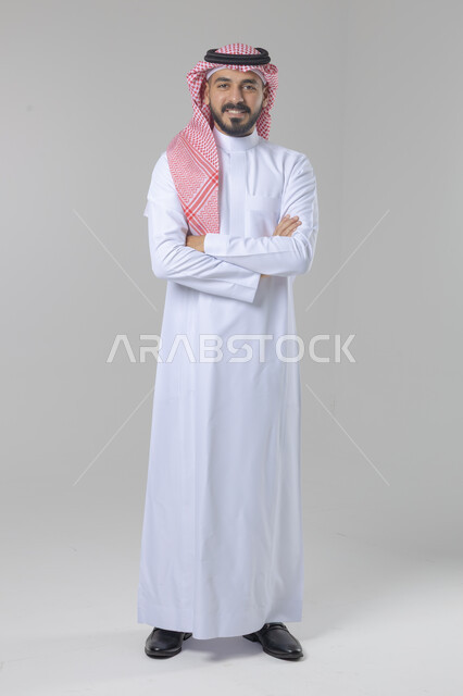 الاعتناء والاهتمام بالمظهر الخارجي، مفهوم الاناقة والرجولة، التقاط صور شخصية للوثائق الرسمية، بورتريه لشاب عربي خليجي سعودي يرتدي الثوب التقليدي والشماغ يقف بأيدي متقاطعة ينظر الى الكاميرا بتعابير السرور والثقة بالنفس، الجسم بالطول الكامل، خلفية بيضاء