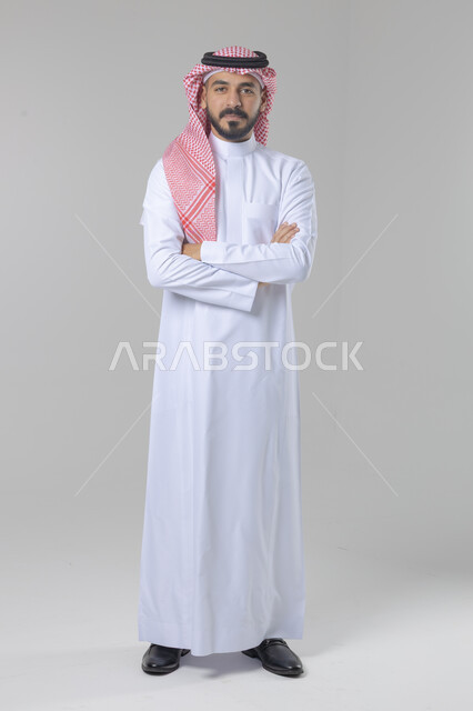 الاعتناء والاهتمام بالمظهر الخارجي، مفهوم الاناقة والرجولة، التقاط صور شخصية للوثائق الرسمية، بورتريه لشاب عربي خليجي سعودي يرتدي الثوب التقليدي والشماغ يقف بأيدي متقاطعة ينظر الى الكاميرا بتعابير السرور والثقة بالنفس، الجسم بالطول الكامل، خلفية بيضاء