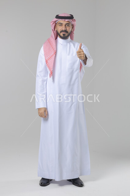  الإمتياز والتميز والنجاح، بورتريه لرجل عربي خليجي سعودي يرتدي الثوب التقليدي والشماغ ويرفع أصابع الإبهام لأعلى، إيماءات السعادة والإعجاب والموافقة، حركة أصبع تدل على القبول والرضا، الجسم بالطول الكامل، خلفية بيضاء 
