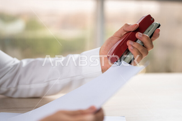 صورة مقربة ليد رجل عربي خليجي سعودي يحمل بيده اوراق خاصة بالعمل، سكرتير يدير الاعمال الورقية، استخدام كابسة الورق، تدبيس الاوراق، مفهوم التوظيف والعمل، أعمال مكتبية