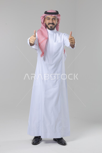  الإمتياز والتميز والنجاح، بورتريه لرجل عربي خليجي سعودي يرتدي الثوب التقليدي والشماغ ويرفع أصابع الإبهام لأعلى، إيماءات السعادة والإعجاب والموافقة، حركة أصبع تدل على القبول والرضا، الجسم بالطول الكامل، خلفية بيضاء 