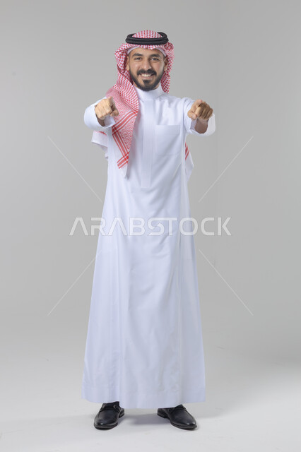 الإشارة إلى شيء ما, الوقوف باستقامة واعتدال, بورتريه لرجل عربي خليجي سعودي يرتدي الثوب التقليدي و الشماغ يشير بيديه لمكان معين, صورة للجسم بالطول الكامل, خلفية بيضاء
