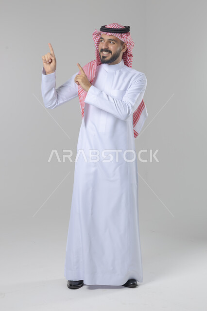 الإشارة إلى شيء ما في الاعلى, الوقوف باستقامة واعتدال, بورتريه لرجل عربي خليجي سعودي يرتدي الثوب التقليدي و الشماغ يشير بيديه لمكان معين, صورة للجسم بالطول الكامل, خلفية بيضاء