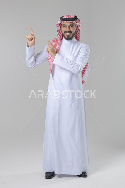 الإشارة إلى شيء ما في الاعلى, الوقوف باستقامة واعتدال, بورتريه لرجل عربي خليجي سعودي يرتدي الثوب التقليدي و الشماغ يشير بيديه لمكان معين, صورة للجسم بالطول الكامل, خلفية بيضاء