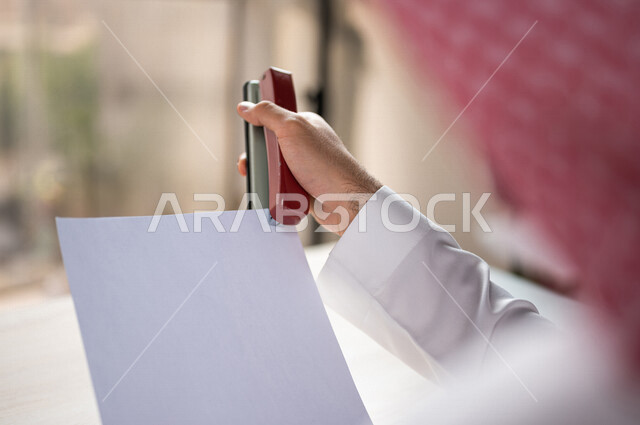 صورة مقربة ليد رجل عربي خليجي سعودي يحمل بيده اوراق خاصة بالعمل، سكرتير يدير الاعمال الورقية، استخدام كابسة الورق، تدبيس الاوراق، مفهوم التوظيف والعمل، أعمال مكتبية