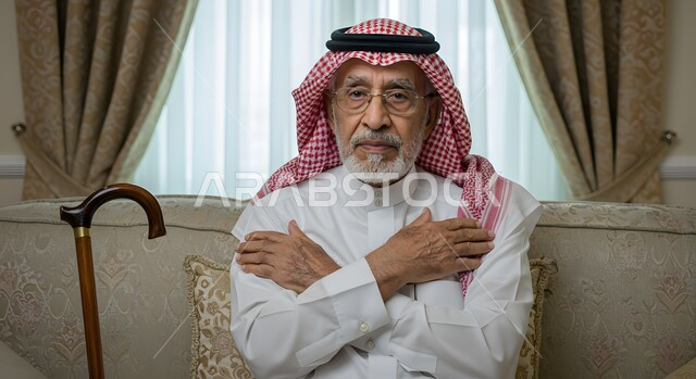 مفهوم الجلوس والراحة، صورة مقربة لرجل عربي خليجي سعودي كبير بالسن مبتسم يرتدي الشماغ والثوب التقليدي يجلس في غرفة المعيشة بجانبه العكاز يضع يديه على كتفيه