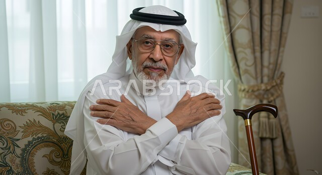مفهوم الجلوس والراحة، صورة مقربة لرجل عربي خليجي سعودي كبير بالسن مبتسم يرتدي الشماغ والثوب التقليدي يجلس في غرفة المعيشة بجانبه العكاز يضع يديه على كتفيه