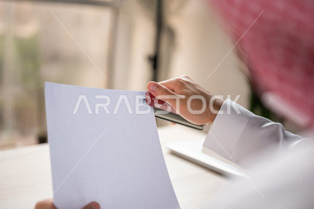 صورة مقربة ليد رجل عربي خليجي سعودي يحمل بيده اوراق خاصة بالعمل، سكرتير يدير الاعمال الورقية، استخدام كابسة الورق، تدبيس الاوراق، مفهوم التوظيف والعمل، أعمال مكتبية