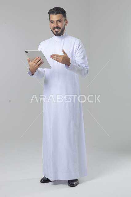 Using modern advanced technology in practical life via tablet, expressions and gestures of integration, portrait of a smiling young Saudi Arabian Gulf man wearing traditional dress working on a tablet, full body image, white background
