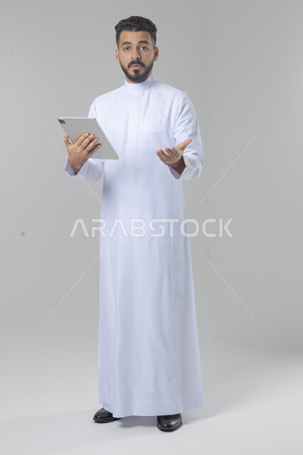 Using modern advanced technology in practical life via tablet, expressions and gestures of integration, portrait of a smiling young Saudi Arabian Gulf man wearing traditional dress working on a tablet, full body image, white background