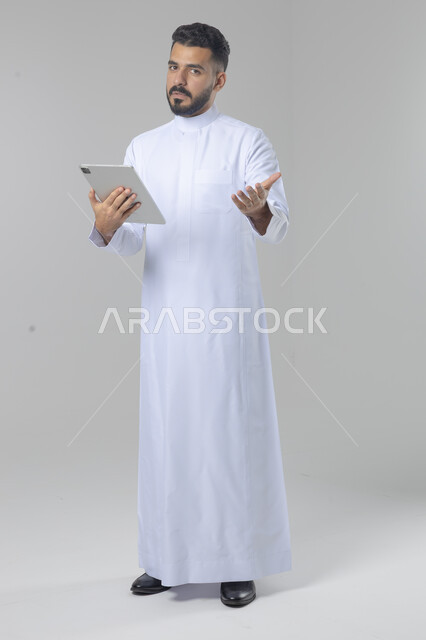 Using modern advanced technology in practical life via tablet, expressions and gestures of integration, portrait of a smiling young Saudi Arabian Gulf man wearing traditional dress working on a tablet, full body image, white background