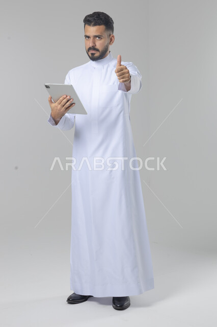 استعمال التقنية الحديثه المتطورة في الحياة العملية عبر الجهاز اللوحي، تعابير وايماءات الاندماج، بورتريه لشاب عربي خليجي سعودي مبتسم يرتدي الثوب التقليدي يقوم بالعمل على جهازالتابلت، صورة الجسم بالطول الكامل، خلفية بيضاء