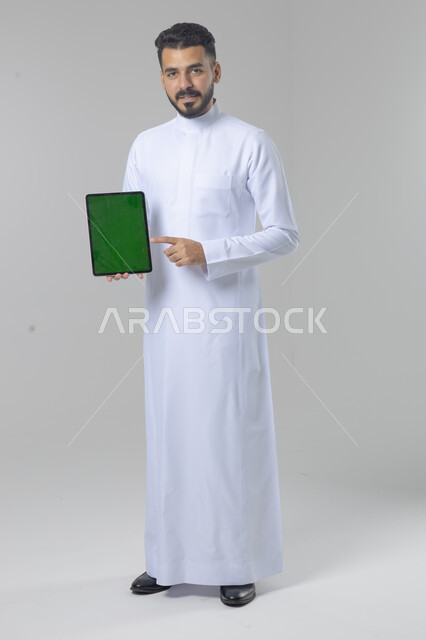 عرض شاشة خضراء كروما عبر الجهاز اللوحي، استعمال التقنية الحديثه المتطورة في الحياة العملية، تعابير وايماءات الاندماج، بورتريه لشاب عربي خليجي سعودي مبتسم يرتدي الثوب التقليدي يقوم بالعمل على جهازالتابلت، صورة الجسم بالطول الكامل، خلفية بيضاء