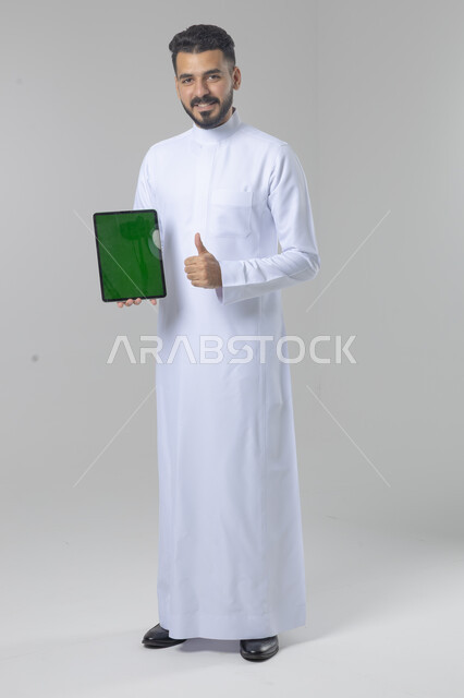 عرض شاشة خضراء كروما عبر الجهاز اللوحي، استعمال التقنية الحديثه المتطورة في الحياة العملية، تعابير وايماءات الاندماج، بورتريه لشاب عربي خليجي سعودي مبتسم يرتدي الثوب التقليدي يقوم بالعمل على جهازالتابلت، صورة الجسم بالطول الكامل، خلفية بيضاء