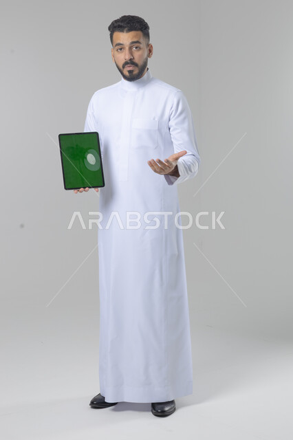 عرض شاشة خضراء كروما عبر الجهاز اللوحي، استعمال التقنية الحديثه المتطورة في الحياة العملية، تعابير وايماءات الاندماج، بورتريه لشاب عربي خليجي سعودي مبتسم يرتدي الثوب التقليدي يقوم بالعمل على جهازالتابلت، صورة الجسم بالطول الكامل، خلفية بيضاء