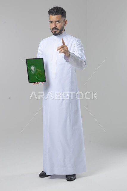 عرض شاشة خضراء كروما عبر الجهاز اللوحي، استعمال التقنية الحديثه المتطورة في الحياة العملية، تعابير وايماءات الاندماج، بورتريه لشاب عربي خليجي سعودي مبتسم يرتدي الثوب التقليدي يقوم بالعمل على جهازالتابلت، صورة الجسم بالطول الكامل، خلفية بيضاء