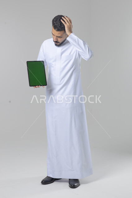 عرض شاشة خضراء كروما عبر الجهاز اللوحي، استعمال التقنية الحديثه المتطورة في الحياة العملية، تعابير وايماءات الاندماج، بورتريه لشاب عربي خليجي سعودي مبتسم يرتدي الثوب التقليدي يقوم بالعمل على جهازالتابلت، صورة الجسم بالطول الكامل، خلفية بيضاء