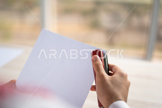 صورة مقربة ليد رجل عربي خليجي سعودي يحمل بيده اوراق خاصة بالعمل، سكرتير يدير الاعمال الورقية، استخدام كابسة الورق، تدبيس الاوراق، مفهوم التوظيف والعمل، أعمال مكتبية