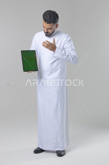 عرض شاشة خضراء كروما عبر الجهاز اللوحي، استعمال التقنية الحديثه المتطورة في الحياة العملية، تعابير وايماءات الاندماج، بورتريه لشاب عربي خليجي سعودي مبتسم يرتدي الثوب التقليدي يقوم بالعمل على جهازالتابلت، صورة الجسم بالطول الكامل، خلفية بيضاء