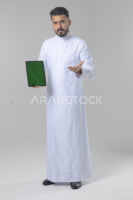 عرض شاشة خضراء كروما عبر الجهاز اللوحي، استعمال التقنية الحديثه المتطورة في الحياة العملية، تعابير وايماءات الاندماج، بورتريه لشاب عربي خليجي سعودي مبتسم يرتدي الثوب التقليدي يقوم بالعمل على جهازالتابلت، صورة الجسم بالطول الكامل، خلفية بيضاء