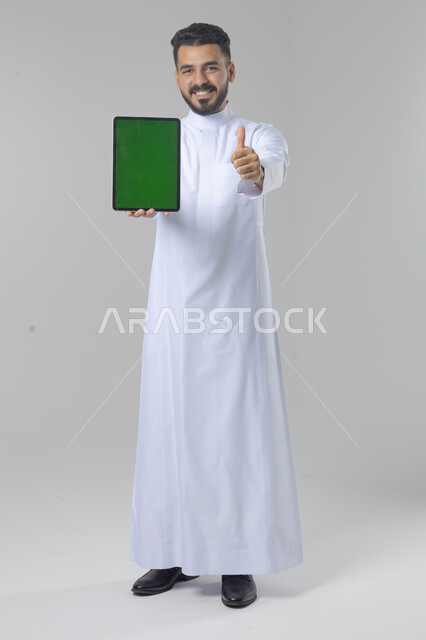 عرض شاشة خضراء كروما عبر الجهاز اللوحي، استعمال التقنية الحديثه المتطورة في الحياة العملية، تعابير وايماءات الاندماج، بورتريه لشاب عربي خليجي سعودي مبتسم يرتدي الثوب التقليدي يقوم بالعمل على جهازالتابلت، صورة الجسم بالطول الكامل، خلفية بيضاء