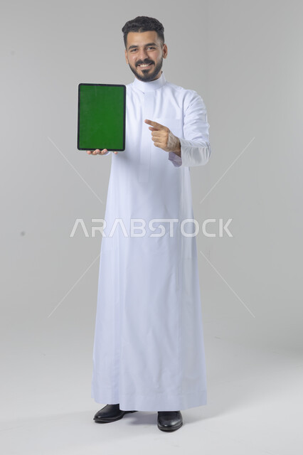 عرض شاشة خضراء كروما عبر الجهاز اللوحي، استعمال التقنية الحديثه المتطورة في الحياة العملية، تعابير وايماءات الاندماج، بورتريه لشاب عربي خليجي سعودي مبتسم يرتدي الثوب التقليدي يقوم بالعمل على جهازالتابلت، صورة الجسم بالطول الكامل، خلفية بيضاء