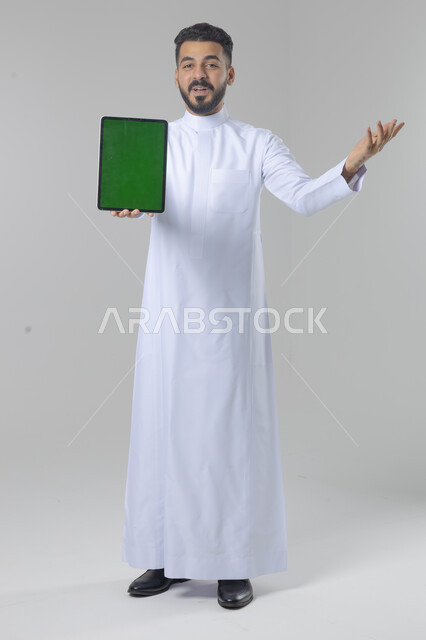 عرض شاشة خضراء كروما عبر الجهاز اللوحي، استعمال التقنية الحديثه المتطورة في الحياة العملية، تعابير وايماءات الاندماج، بورتريه لشاب عربي خليجي سعودي مبتسم يرتدي الثوب التقليدي يقوم بالعمل على جهازالتابلت، صورة الجسم بالطول الكامل، خلفية بيضاء