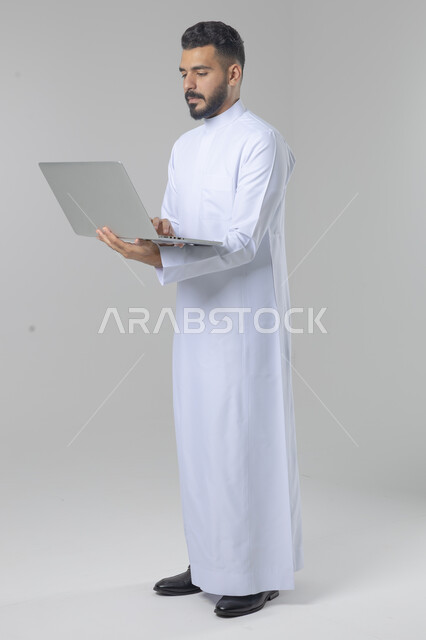 استخدام التقنية الحديثه المتطورة في الحياة العملية، تعابير وايماءات الاندماج والسرور، بورتريه لشاب عربي خليجي سعودي مبتسم يرتدي الثوب التقليدي يقوم بالعمل على الحاسب المحمول، صورة الجسم بالطول الكامل، خلفية بيضاء