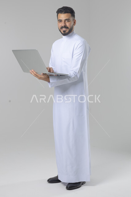 استخدام التقنية الحديثه المتطورة في الحياة العملية، تعابير وايماءات الاندماج والسرور، بورتريه لشاب عربي خليجي سعودي مبتسم يرتدي الثوب التقليدي يقوم بالعمل على الحاسب المحمول، صورة الجسم بالطول الكامل، خلفية بيضاء