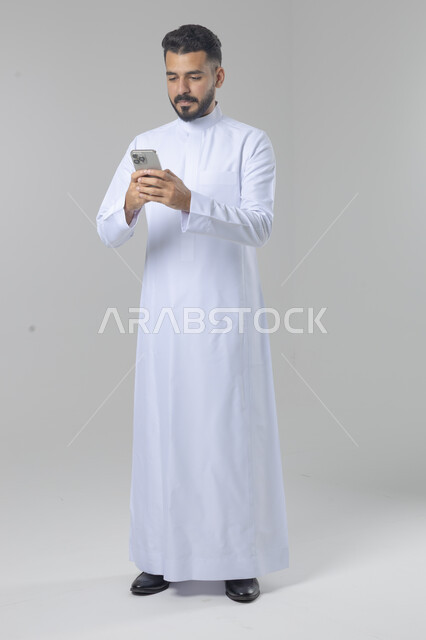  تعابير وايماءات الاندماج والسرور، استخدام التقنية الحديثه المتطورة في الحياة العملية، بورتريه لشاب عربي خليجي سعودي مبتسم يرتدي الثوب التقليدي يقوم بالعمل على الهاتف المحمول، صورة الجسم بالطول الكامل، خلفية بيضاء