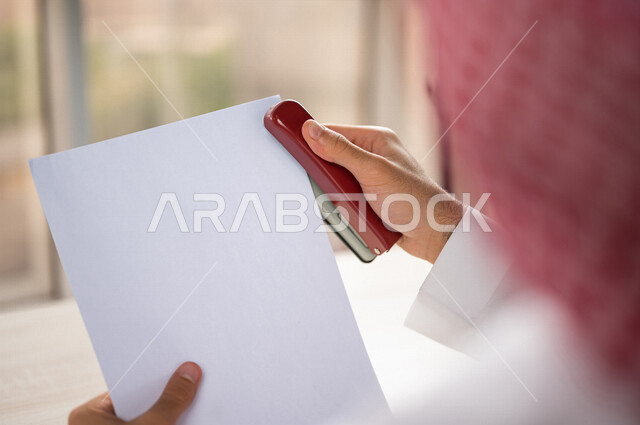 صورة مقربة ليد رجل عربي خليجي سعودي يحمل بيده اوراق خاصة بالعمل، سكرتير يدير الاعمال الورقية، استخدام كابسة الورق، تدبيس الاوراق، مفهوم التوظيف والعمل، أعمال مكتبية