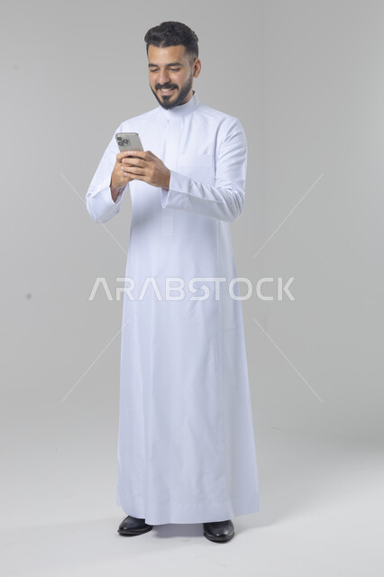  تعابير وايماءات الاندماج والسرور، استخدام التقنية الحديثه المتطورة في الحياة العملية، بورتريه لشاب عربي خليجي سعودي مبتسم يرتدي الثوب التقليدي يقوم بالعمل على الهاتف المحمول، صورة الجسم بالطول الكامل، خلفية بيضاء
