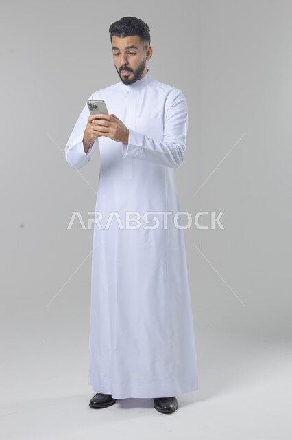  تعابير وايماءات الاندماج والسرور، استخدام التقنية الحديثه المتطورة في الحياة العملية، بورتريه لشاب عربي خليجي سعودي مبتسم يرتدي الثوب التقليدي يقوم بالعمل على الهاتف المحمول، صورة الجسم بالطول الكامل، خلفية بيضاء