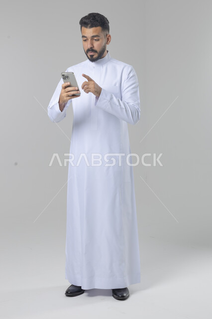  تعابير وايماءات الاندماج والسرور، استخدام التقنية الحديثه المتطورة في الحياة العملية، بورتريه لشاب عربي خليجي سعودي مبتسم يرتدي الثوب التقليدي يقوم بالعمل على الهاتف المحمول، صورة الجسم بالطول الكامل، خلفية بيضاء