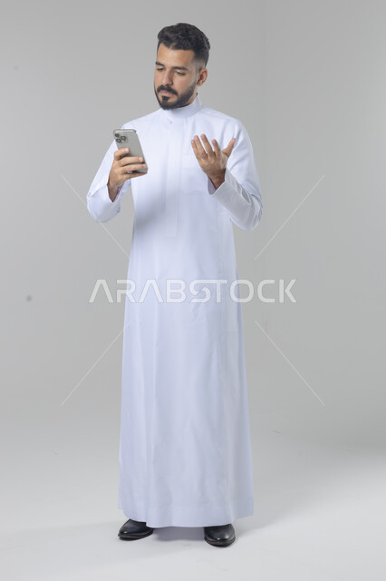  تعابير وايماءات الاندماج والسرور، استخدام التقنية الحديثه المتطورة في الحياة العملية، بورتريه لشاب عربي خليجي سعودي مبتسم يرتدي الثوب التقليدي يقوم بالعمل على الهاتف المحمول، صورة الجسم بالطول الكامل، خلفية بيضاء