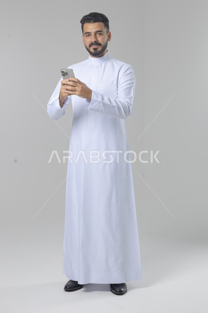  تعابير وايماءات الاندماج والسرور، استخدام التقنية الحديثه المتطورة في الحياة العملية، بورتريه لشاب عربي خليجي سعودي مبتسم يرتدي الثوب التقليدي يقوم بالعمل على الهاتف المحمول، صورة الجسم بالطول الكامل، خلفية بيضاء