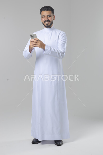  تعابير وايماءات الاندماج والسرور، استخدام التقنية الحديثه المتطورة في الحياة العملية، بورتريه لشاب عربي خليجي سعودي مبتسم يرتدي الثوب التقليدي يقوم بالعمل على الهاتف المحمول، صورة الجسم بالطول الكامل، خلفية بيضاء