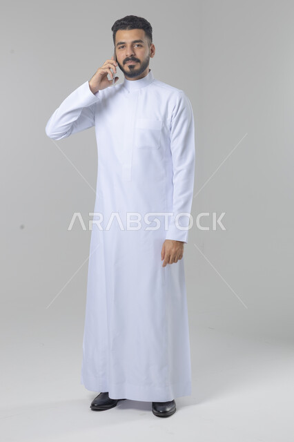 Chatting and conversation using a modern technical device, answering a phone call, portrait of a Saudi Arabian Gulf man wearing traditional thobe making a phone call through a mobile phone, full body image, white background