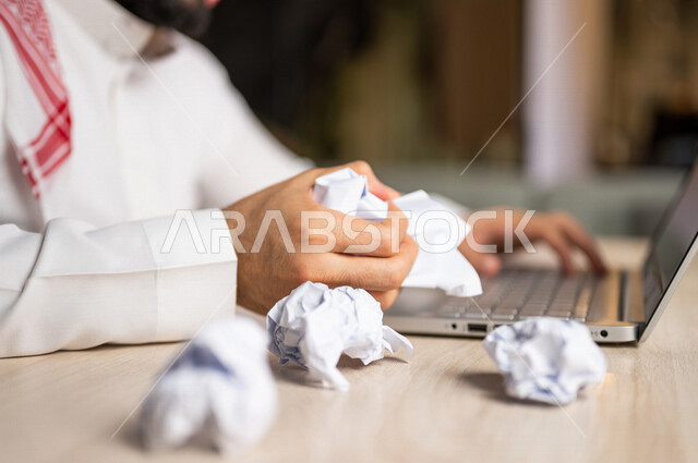 A Saudi Arabian Gulf man sitting in front of a laptop, creasing papers, searching for ideas and plans, failing to write and blogging, failing to be creative and working, trying to find solutions