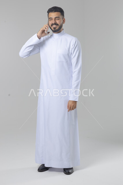 Chatting and conversation using a modern technical device, answering a phone call, portrait of a Saudi Arabian Gulf man wearing traditional thobe making a phone call through a mobile phone, full body image, white background
