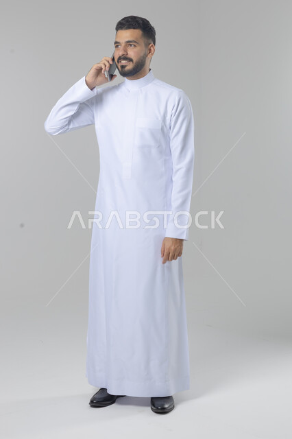 الدردشة والمحادثة باستخدام جهاز تقني حديث, الرد على مكالمة هاتفية، بورتريه لرجل عربي خليجي سعودي يرتدي الثوب التقليدي يقوم باجراء اتصال هاتفي من خلال الهاتف المحمول، صورة الجسم بالطول الكامل, خلفية بيضاء