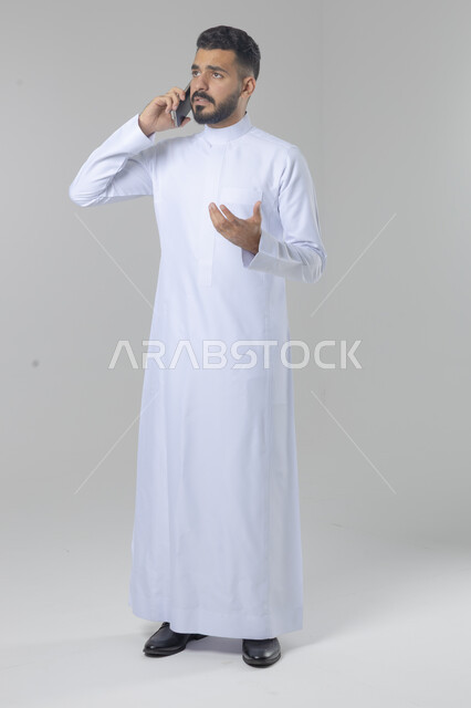 الدردشة والمحادثة باستخدام جهاز تقني حديث, الرد على مكالمة هاتفية، بورتريه لرجل عربي خليجي سعودي يرتدي الثوب التقليدي يقوم باجراء اتصال هاتفي من خلال الهاتف المحمول، صورة الجسم بالطول الكامل, خلفية بيضاء