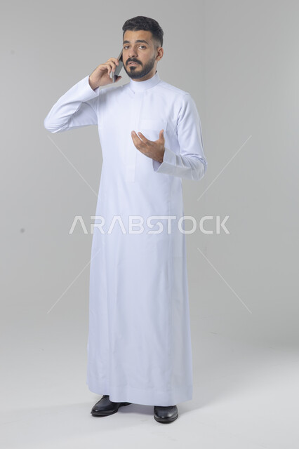 الدردشة والمحادثة باستخدام جهاز تقني حديث, الرد على مكالمة هاتفية، بورتريه لرجل عربي خليجي سعودي يرتدي الثوب التقليدي يقوم باجراء اتصال هاتفي من خلال الهاتف المحمول، صورة الجسم بالطول الكامل, خلفية بيضاء