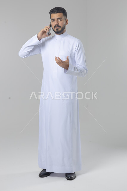 الدردشة والمحادثة باستخدام جهاز تقني حديث, الرد على مكالمة هاتفية، بورتريه لرجل عربي خليجي سعودي يرتدي الثوب التقليدي يقوم باجراء اتصال هاتفي من خلال الهاتف المحمول، صورة الجسم بالطول الكامل, خلفية بيضاء