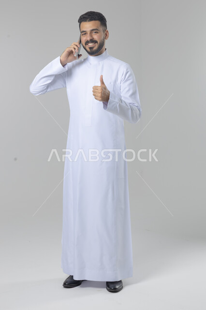 Chatting and conversation using a modern technical device, answering a phone call, portrait of a Saudi Arabian Gulf man wearing traditional thobe making a phone call through a mobile phone, full body image, white background