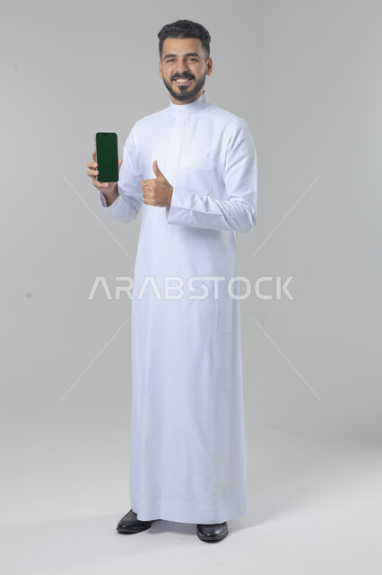  تعابير وايماءات الاندماج والسرور، استخدام التقنية الحديثه المتطورة في الحياة العملية، بورتريه لشاب عربي خليجي سعودي مبتسم يرتدي الثوب التقليدي يقوم بعرض شاشة فارغة على الهاتف المحمول، صورة الجسم بالطول الكامل، خلفية بيضاء